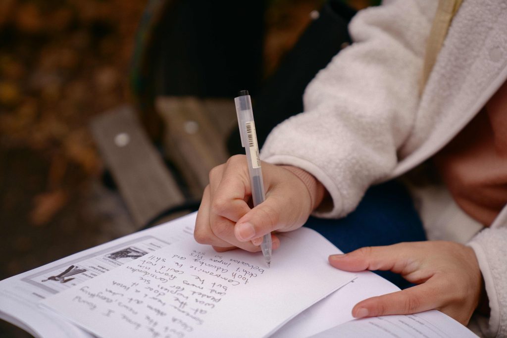 A woman seated on the bench and writing, with fall leaves on the ground.